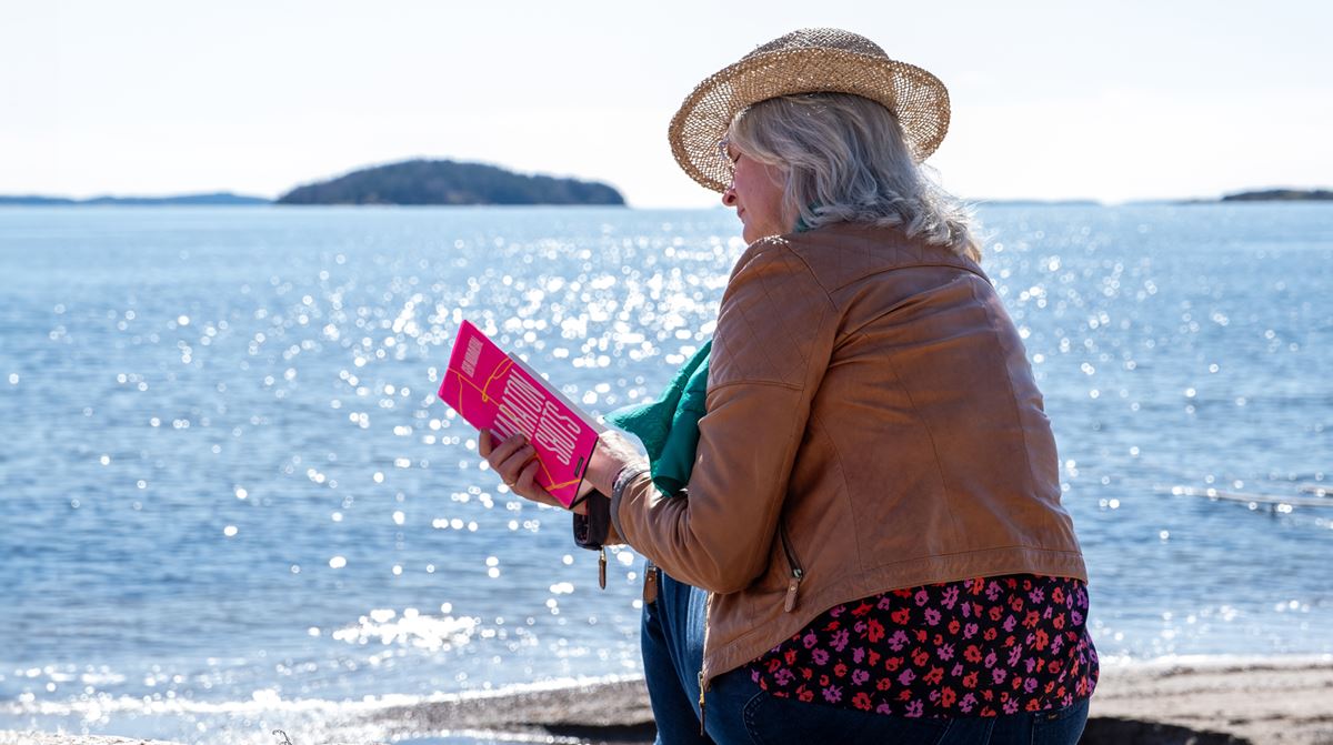 Dame med hatt leser bok ved sjøen en godværsdag.